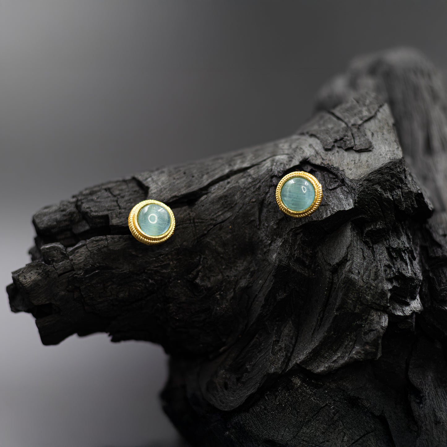 DIVAMAS Aqua blue Monalisa stone stud earrings with a shimmering finish and gold bezel detailing, placed against a dark backdrop